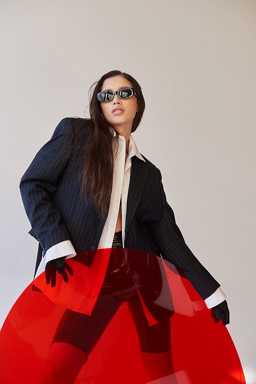 edgy style, studio photography, young asian model in stylish look and sunglasses posing near red round shaped glass, grey background, blazer and latex shorts, youthful fashion, modern woman