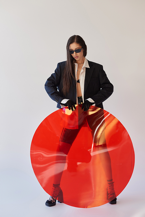 attractive asian model in stylish look and sunglasses posing holding red round shaped glass, grey background, blazer and latex shorts, youthful and modern woman, fashion forward, studio photography