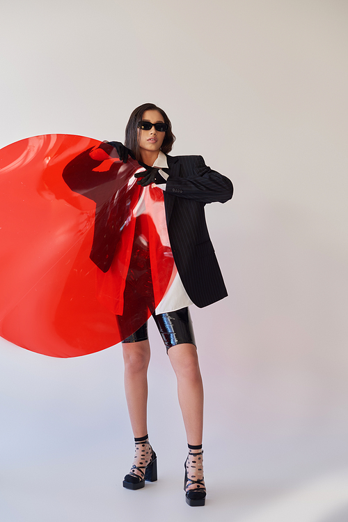 pretty asian model in stylish look and sunglasses posing holding red round shaped glass, grey background, blazer and latex shorts, youthful and modern woman, fashion statement, studio photography