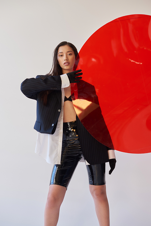 beautiful asian woman in trendy outfit holding red round shaped glass, grey background, blazer and black latex shorts, youthful model in gloves, fashion forward, studio photography, conceptual