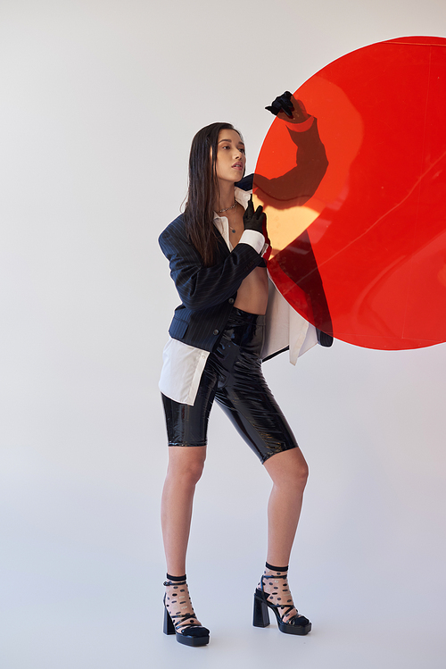 pretty asian woman in trendy outfit holding red round shaped glass, grey background, blazer and black latex shorts, youthful model in gloves, fashion and style, studio photography, conceptual