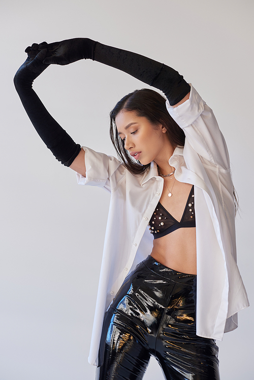 personal style, young asian woman with brunette hair posing with raised hands on grey background, white shirt and black gloves, model posing in latex shorts in studio, fashion statement, bold style