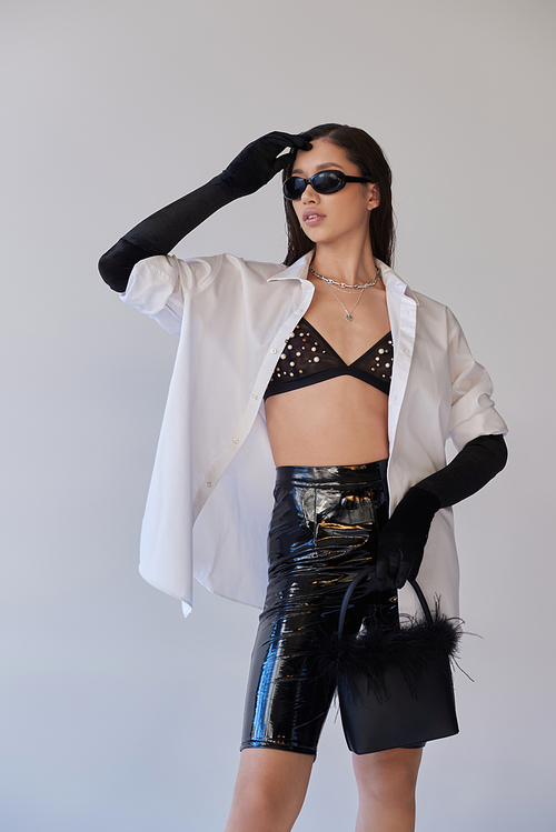 trendy look, fashion forward, brunette asian woman in sunglasses posing with feathered purse on grey background, young model in latex shorts, black gloves and white shirt, conceptual