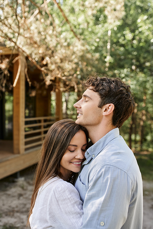 Smiling couple with closed eyes embracing and standing near vacation house at background outdoors