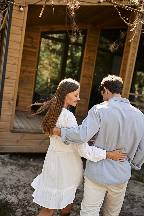 Smiling brunette woman in sundress hugging boyfriend and walking near summer house outdoors