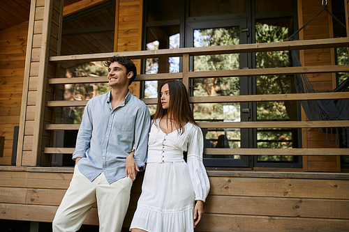 Smiling man in casual clothes standing near girlfriend in sundress and vacation house at background