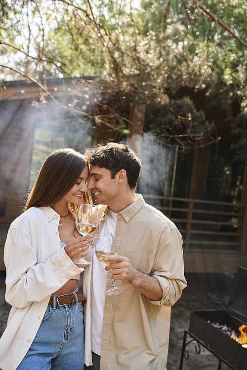 Smiling brunette couple in casual clothes holding wine during picnic near vacation house outdoors