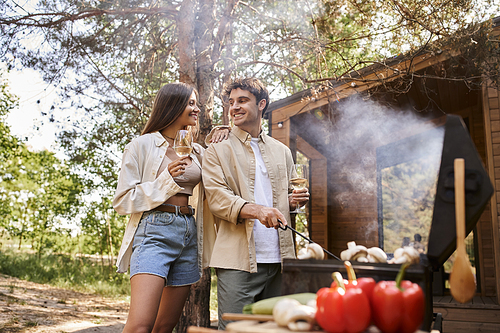 Smiling woman holding wine and standing near boyfriend, bbq with vacation house at background