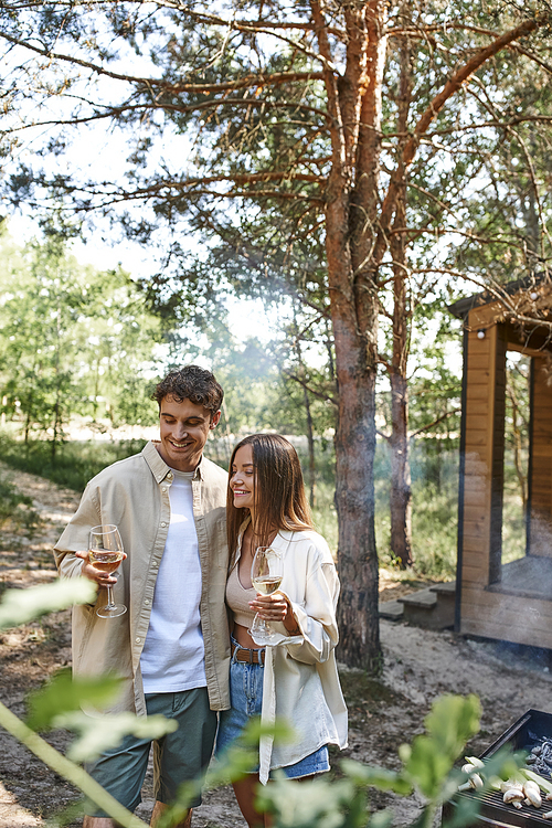 Joyful couple in casual clothes holding wine near barbecue and blurred vacation house at background