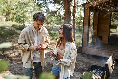 Smiling woman holding wine and talking to boyfriend near grill and vacation house at background