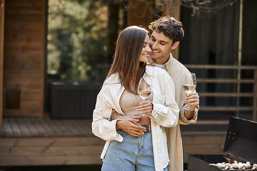 Smiling man hugging girlfriend and holding wine near bbq and blurred vacation house at background
