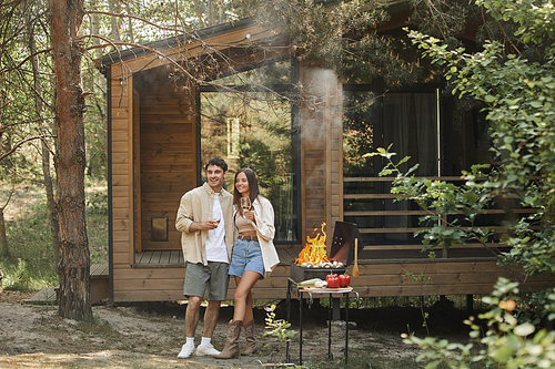 Positive romantic couple holding wine and standing near grill and vacation house at background