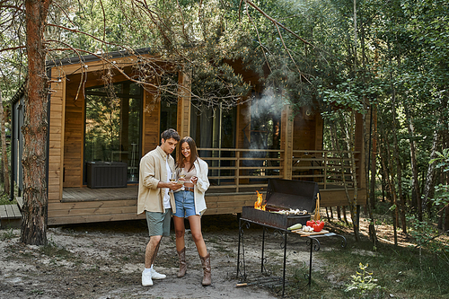 Smiling romantic couple toasting with wine and standing near barbecue and vacation house outdoors