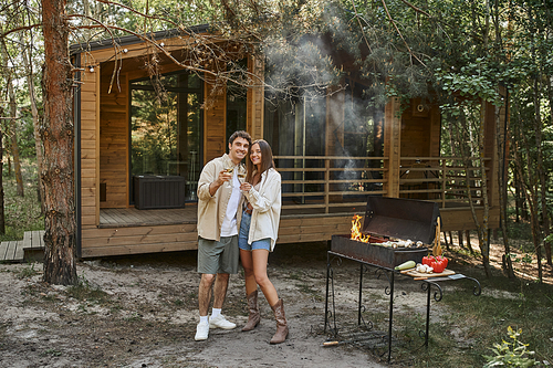 Positive romantic couple holding wine and looking at camera near grill and vacation house