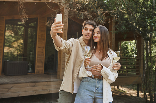 Stylish man taking selfie on smartphone with girlfriend holding wine near summer house at background