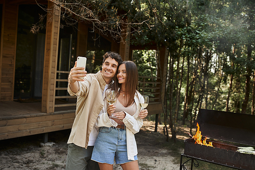 Smiling couple with wine taking selfie on smartphone near bbq and vacation house at background