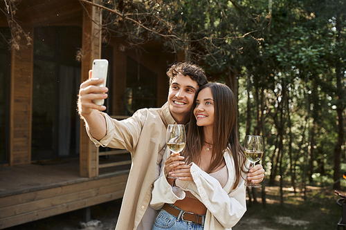 Smiling young woman holding wine while boyfriend taking selfie on smartphone near vacation house