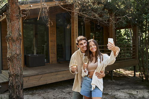 Smiling man hugging girlfriend and taking selfie with girlfriend holding wine near vacation house