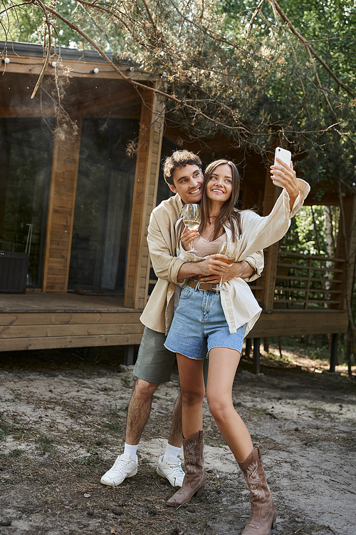 Smiling man hugging girlfriend with wine taking selfie on smartphone near vacation house outdoors