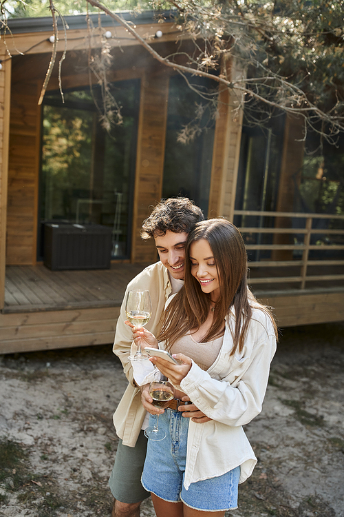 Smiling and stylish couple with wine using mobile phone near blurred vacation house at background