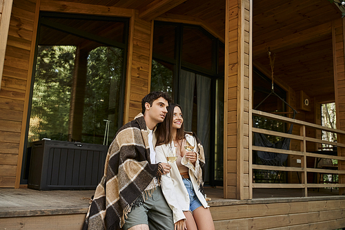 Smiling couple in blanket holding wine and looking away on porch of summer house