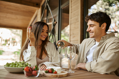 Smiling man pouring wine near girlfriend with strawberry on terrace of vacation house at background