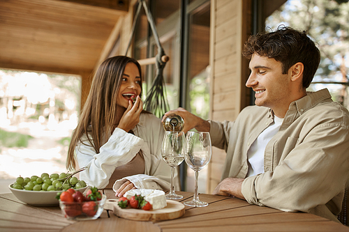 Smiling man pouring wine while girlfriend eating strawberry on terrace of vacation house