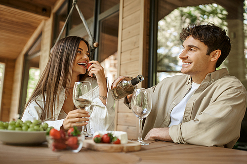 Smiling woman eating strawberry near boyfriend with wine on terrace of wooden vacation house