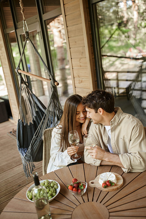 Positive woman hugging boyfriend near wine and food on terrace of wooden summer house
