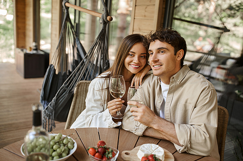 Happy romantic couple holding wine near fruits and cheese on terrace of vacation house during summer