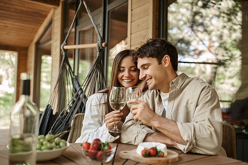 Smiling romantic couple clinking with wine near blurred food on terrace of wooden vacation house