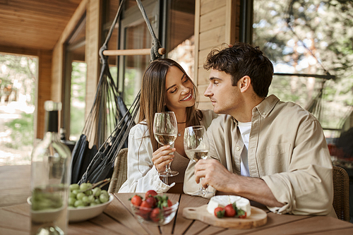 Smiling woman holding wine and looking at boyfriend near blurred food on terrace of summer house