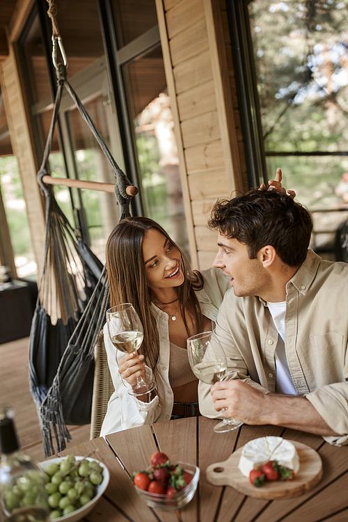 Smiling woman with wine talking and touching boyfriend near food on terrace of vacation house