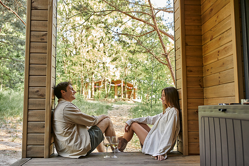 Side view of positive romantic couple sitting near wine on porch of wooden summer house outdoors