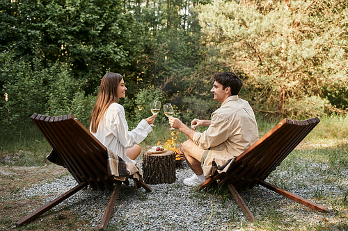 Side view of smiling romantic couple holding wine near food and firewood during picnic outdoors