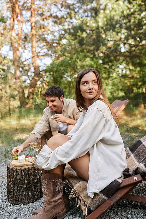 Brunette woman sitting on deck chair near boyfriend with wine and food during picnic outdoors