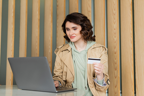 pleased woman with wavy brunette hair, in beige trench coat holding credit card and looking at laptop while sitting at table in lobby of contemporary hotel during online shopping, leisure and travel