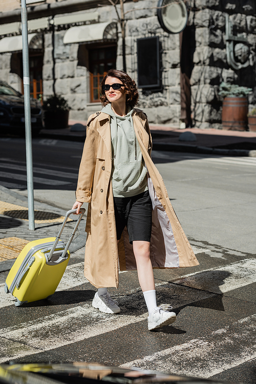 full length of self-assured and independent woman in beige trench coat, grey hoodie and black shorts holding yellow suitcase while crossing road in European city, urban lifestyle