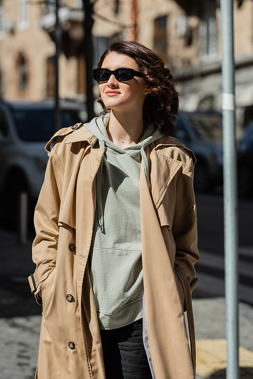 young and positive woman with wavy brunette hair, in dark sunglasses, grey hoodie and beige trench coat looking away on blurred background in European city, street photography