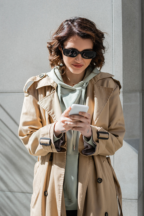 young and carefree woman with wavy brunette hair and tattoo, in dark sunglasses, beige trench coat and hoodie browsing internet on mobile phone while standing near grey wall in sunlight on street