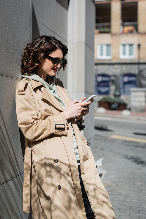 cheerful and trendy woman with wavy brunette hair and tattoo, in dark sunglasses and beige trench coat standing near grey building and browsing internet on mobile phone on street in european city