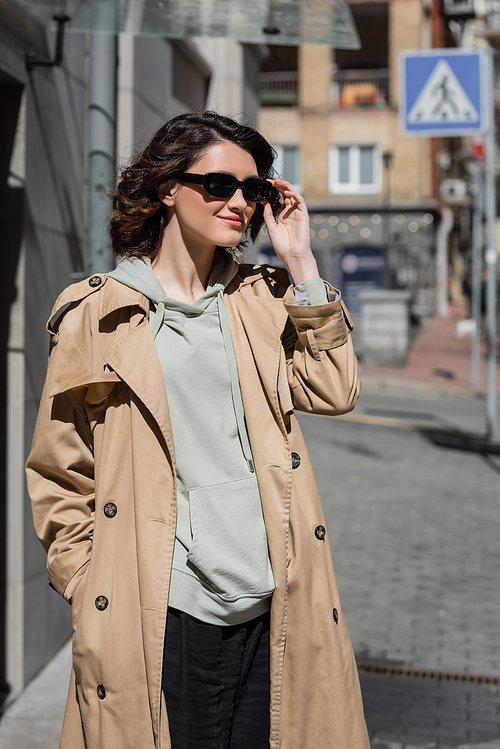 young appealing woman with wavy brunette hair adjusting dark stylish sunglasses while standing in grey hoodie and beige trench coat in city on blurred background, street photography