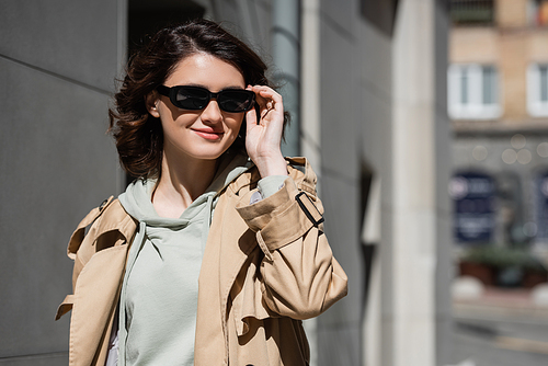 young and pleased woman with wavy brunette hair, in grey hoodie and beige trench coat adjusting dark stylish sunglasses and looking away on urban street on blurred background