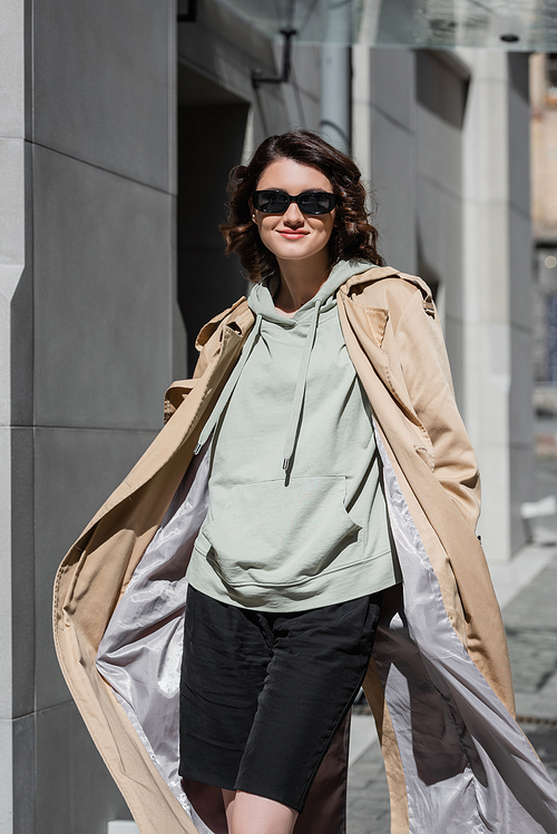 stylish and carefree woman in dark sunglasses, grey hoodie and beige trench coat posing on street in modern European city and looking at camera, travel lifestyle, urban fashion