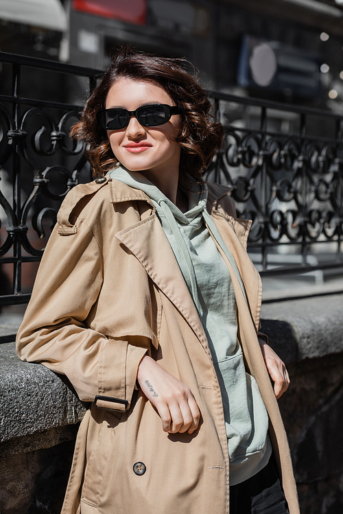 charismatic tattooed woman with wavy brunette hair, in dark stylish sunglasses, grey hoodie and beige trench coat smiling and looking away near forged fence in city, street photography