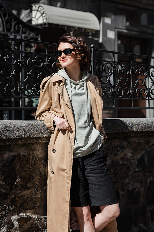 travel lifestyle, trendy and cheerful woman in grey hoodie, beige trench coat and stylish sunglasses standing with hand in pocket of black shorts and looking away near forged fence on city street