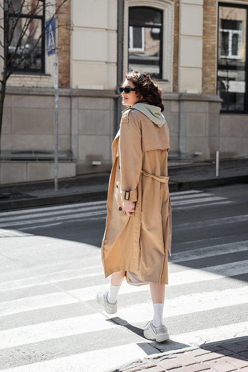 full length of fashionable woman in grey hoodie, beige trench coat and white sneakers crossing road and looking away in European city, travel lifestyle, urban fashion