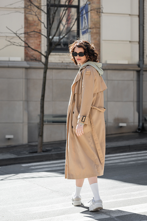 full length of young and stylish woman with wavy brunette hair crossing road in dark sunglasses, grey hoodie, beige trench coat and white sneakers and looking at camera in European city