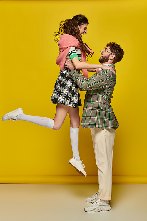 joyful couple having fun, man in glasses lifting excited young woman on yellow backdrop, students