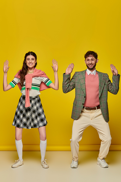 funny couple, happy young man and woman gesturing, standing on yellow backdrop, looking at camera